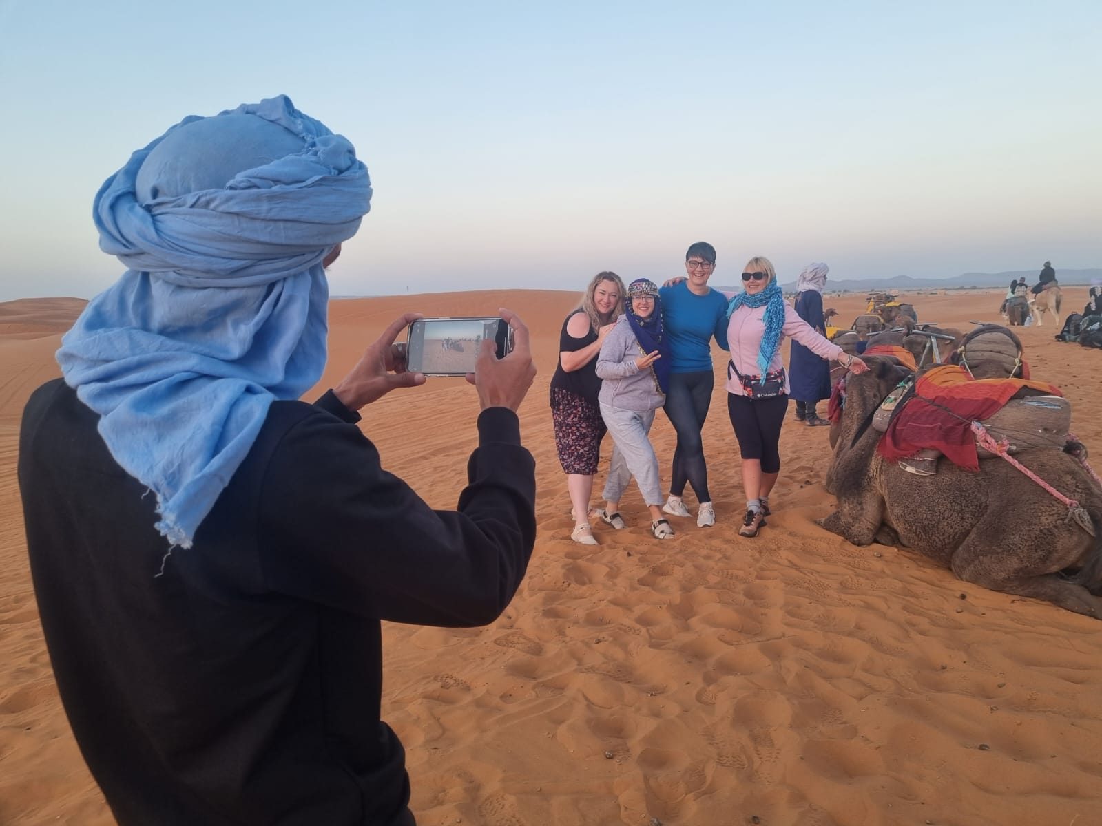 Man in a blue turban photographing a group beside resting camels in the dunes