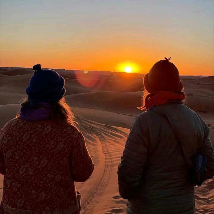 Two people watching the sun rise over desert dunes